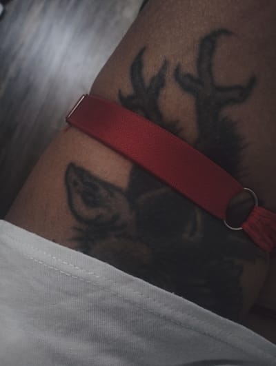 A close-up shot shows a person's upper thigh with a tattoo of a wolf and antlers, partially obscured by a red strap with a metal loop. The person is wearing a white garment at the bottom of the frame. The location is not discernible from the image.