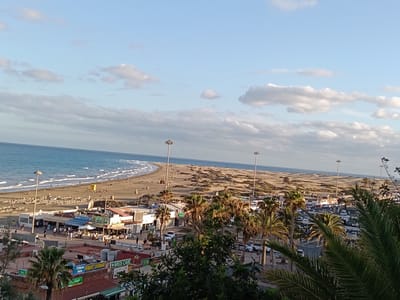 The image shows a coastal scene in Playa del Inglés, Spain. In the foreground are buildings with shops and restaurants along a walkway where people stroll and dine. Palm trees are scattered throughout this area. Beyond this, there is a wide, sandy beach with rows of sun loungers and a lifeguard station. People are walking along the shoreline and enjoying the ocean. In the distance, there are sand dunes, adding to the desert-like landscape that characterizes parts of Gran Canaria. Overhead lighting posts line the scene. Above, the sky is blue with scattered white clouds. The ocean extends to the horizon, meeting the sky.