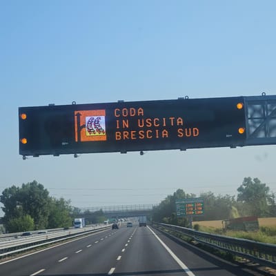 This media file captures a highway scene in San Zeno Naviglio, Italy, on a clear, sunny day. The primary focus is an overhead electronic traffic sign displaying a message in orange LED lights. The sign reads "CODA IN USCITA BRESCIA SUD," which translates to "Traffic Jam at Exit Brescia South." To the left of the text, a graphic depicts a queue of cars, further reinforcing the message of congestion.

Below the traffic sign, another electronic display provides fuel prices at various distances. It lists "prezzo carburanti self-service" (self-service fuel prices) and shows prices for "benzina" (gasoline) and "gasolio" (diesel) at 4.6 km, 40 km, and 69 km distances. For instance, at 4.6 km, both gasoline and diesel are priced at €2.089 per liter.

The highway itself is visible, with multiple lanes and standard white lane markings. Cars and trucks are driving on the road, though the traffic sign suggests potential delays ahead. The surrounding environment features lush green trees and vegetation, with a field of crops and agricultural machinery, including a tractor with a trailer, visible to the right of the highway. The bright sunlight and clear blue sky indicate fair weather. The overall impression is one of normal highway travel interrupted by a traffic issue, with practical information about fuel prices also being provided to drivers.