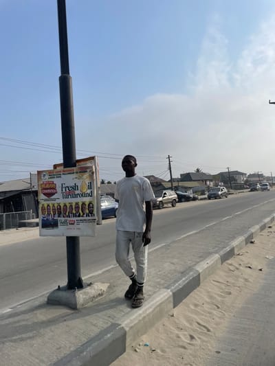 A man stands on a roadside next to a street lamp post with advertisements attached. The man is wearing a white shirt and light-colored pants. Cars are parked along the side of the road in the background. The sky is partially cloudy, and the area appears to be a mix of residential and commercial buildings.
