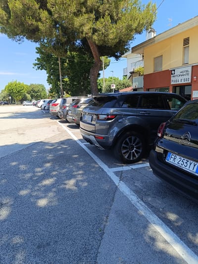 In the city of Riccione, Italy, a parking lot is depicted on a sunny day. Several cars are parked in diagonal spaces marked by white lines. The foreground shows the rear of a black Citroen car with license plate "FR 253YY" and the word "CITROËN" visible. To its left, a dark grey Range Rover Evoque is parked, with its rear tire and part of its body visible. Further down the row, other vehicles, including SUVs and sedans, are parked. Behind the cars, a building with a yellow facade and a dark red lower section is present. A sign on the building reads "HOTEL RISTORANTE PIADA D'ORO". A large tree with green foliage shades a portion of the parking lot. The sky is clear and blue. The ground surface is asphalt, with some patches of dirt and scattered leaves. The overall scene is a typical outdoor parking area with vehicles and surrounding infrastructure under bright daylight.