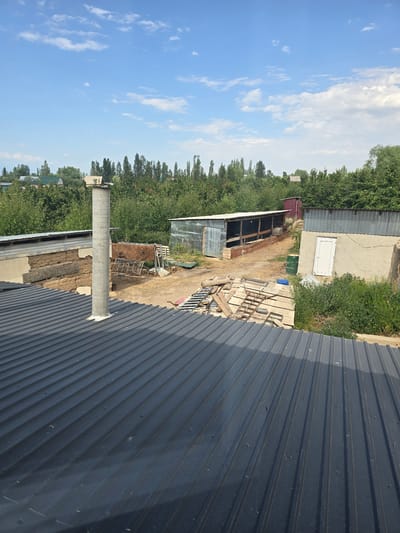 A sunny, daytime, high-angle exterior shot captures a rural or semi-rural area with agricultural buildings and lush green trees under a bright blue sky with scattered white clouds. In the foreground, a dark gray corrugated metal roof slopes downwards. A tall, cylindrical concrete chimney emerges from the roof. Beyond the roof, a light-colored building with a white door is visible, adjacent to a long, low structure with a corrugated metal roof and open sides, appearing to be an animal shelter or storage shed. A pile of wood and metal scraps is present in the dirt yard between these structures. In the background, a dense line of trees forms a backdrop, with hints of houses and more trees in the distance. The scene is located in Chychkan, Kyrgyzstan.
