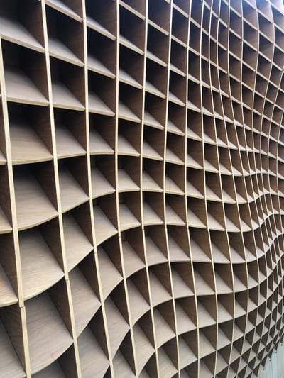 The image shows a detailed, close-up perspective of an architectural feature, specifically an undulating wooden grid structure. The grid is composed of light-colored, presumably wooden, slats forming numerous rectangular or near-rectangular cells. These cells are not uniform in size or shape due to the overall wave-like design of the structure. The structure seems to be viewed from a low angle, creating a sense of depth and perspective that emphasizes the curves and contours. The wooden material appears to have a visible grain and texture, adding a tactile element to the visual information.
This wooden grid is a distinctive feature of the Bolsover Library and Learning Centre in Bolsover, United Kingdom.
