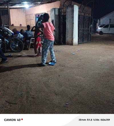 The image depicts an outdoor night scene in Egede, Nigeria, illuminated by artificial light. In the foreground, a young individual, seen from behind, wears a light pink t-shirt and grey patterned trousers, with arms raised in a gesture suggesting dancing or movement. A blue plastic wrapper lies on the unpaved, reddish-brown dirt ground near their feet, which is scattered with small debris. To the immediate left of this individual, another person in a red patterned dress is partially visible.

In the background on the left, a group of approximately 5-6 individuals are seated and standing in front of a light-colored building with a corrugated roof and a barred window. One man, wearing a grey t-shirt and a cap, is seated prominently. A motorcycle is partially visible on the far left. To the right of the central figures, a concrete pillar frames an ornate, dark metal gate, which appears to be part of a compound entrance. Beyond the gate on the far right, two white cars are parked in front of another light-colored building. The ambient lighting suggests multiple light sources illuminating the immediate area.
