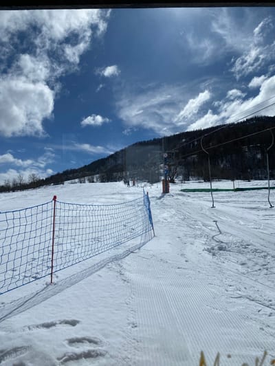 The image depicts a snow-covered ski slope in La Salle-les-Alpes, France, under a partially cloudy blue sky. A blue plastic safety net with red support poles extends across the lower left and mid-ground, casting distinct shadows on the snow. The snow in the foreground shows parallel grooming patterns, while areas near the net display footprints. In the mid-distance, several indistinct figures are visible on the slope, appearing to move towards the base of a ski lift. The ski lift system includes dark pylons and cables ascending the mountain, with some empty lift chairs or T-bars hanging. Orange safety markers are also present near the lift's base. The background consists of a snow-dusted mountain slope densely forested with dark trees. The scene is illuminated by bright sunlight, and the image exhibits reflections or smudges consistent with being photographed through a window or clear barrier, with a dark bar at the top and a faint yellow-orange blur at the bottom right.