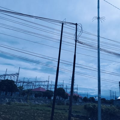 The image displays an outdoor scene dominated by electrical infrastructure under an overcast, blue-hued sky. In the foreground, numerous dark power lines stretch horizontally across the frame, supported by several utility poles. Two prominent dark poles stand near the center, with one showing coiled wires and another a triangular wire support. To the right, a lighter-colored pole features a distinct spider-like metal apparatus attached to it.
The midground shows a patch of green grass or field, bordered by a low concrete wall. Behind this wall, several dark-leaved trees are visible, along with multiple houses characterized by their distinctive red-tiled roofs. In the background, a large electricity substation or power plant extends across the horizon, consisting of a complex array of tall, metallic structures and transmission towers.
The overall color palette is cool and desaturated, indicative of either an overcast day or a specific photographic filter. The heavy cloud cover suggests a subdued, potentially rainy or pre-rainy atmosphere. The scene is static, with no human presence or activities visible. The setting points to an industrialized or semi-urban area, consistent with Teungoh Baro, Indonesia. No prominent text is visible within the main image.