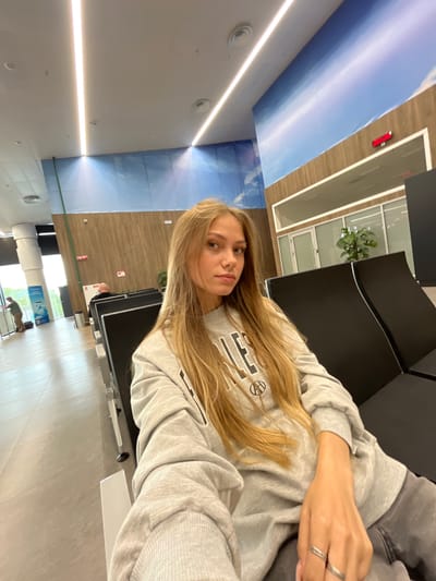A young woman with long blonde hair is sitting in a waiting area, possibly an airport or train station, in Старое Мартьяново, Russia. She is wearing a grey sweatshirt and dark pants, and is looking towards the camera. The waiting area has rows of black chairs and a large mural on the wall depicting a sky scene. There are other people in the background, as well as some signs and potted plants. The lighting in the area is bright, with overhead fluorescent lights.