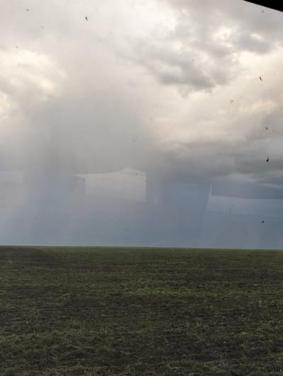 The image depicts an overcast sky with dark, heavy clouds over a flat, open field. The foreground is a cultivated field with dark soil and some scattered green vegetation. In the middle ground, there is a hazy or reflective element that obscures the distant landscape, showing indistinct shapes that could be buildings or natural formations. The overall impression is one of a rural or agricultural setting under a dramatic sky. The provided context of Titov, Kazakhstan, suggests a possible location for this type of landscape.