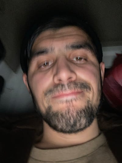 The image displays a close-up, front-facing portrait of an adult male, estimated to be between 20 and 30 years old. He has dark, short hair, prominent dark eyebrows, and brown eyes. A full beard and mustache are present on his face. His expression is characterized by a slight smile. He is dressed in a light brown or tan crew-neck shirt. The background is dark, out of focus, and largely indistinct, suggesting a low-light indoor setting. A blurred red object is partially visible in the upper right quadrant, and a dark, amorphous shape occupies the left side. The primary illumination source appears to be directed at the subject's face. The specific location within the city of Chilаn, Uzbekistan, cannot be determined from the visual information provided in the image.