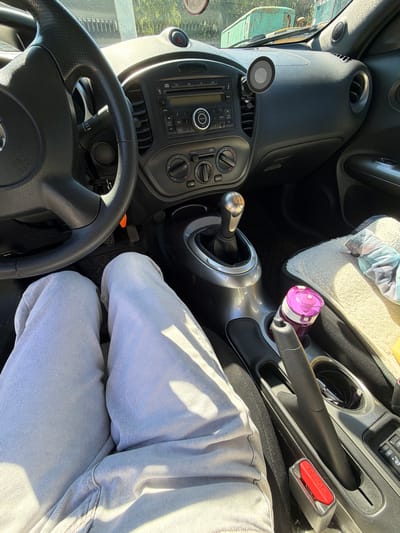 The image shows the interior of a car, viewed from the perspective of the driver. The driver's legs, clad in light-colored jeans, are visible in the lower portion of the frame. The steering wheel, dashboard, gear stick, and center console are prominently featured. A purple water bottle is in a cup holder, and a fluffy blanket is draped over the passenger seat. The car appears to be parked, possibly in a rural or suburban setting, as evidenced by the blurry background which includes a large green structure and trees. There is no indication of the specific location within the city of Ягул, Russia, but the general scene is consistent with a vehicle parked outdoors.