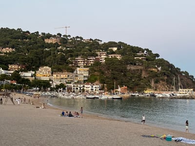 The image captures a seaside town scene in Llafranc, Spain, likely during the late afternoon or early evening as indicated by the soft lighting. In the foreground, a sandy beach is sparsely populated with individuals and small groups. Some people are seated on towels, others are standing or walking along the shoreline. A lifeguard tower with a ladder is visible on the left side of the beach.
The middle ground features a calm body of water, likely the sea, where numerous boats are moored in a harbor. These boats range in size and type, with white hulls and masts dominating the view. A breakwater or pier, constructed of rocks, extends into the water on the right side of the harbor.
Behind the harbor, the town of Llafranc is built into a lush, green hillside. Buildings, primarily hotels or apartment complexes, are clustered together, many with balconies and illuminated windows. The architecture appears modern, with white facades and darker window frames. Dense trees and vegetation cover the slopes between and around the buildings, creating a vibrant green backdrop. A construction crane is visible on the highest point of the hill, suggesting ongoing development. The sky above is a pale, hazy blue, typical of dusk.