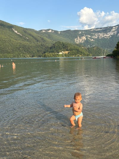 In this vertical, natural light photograph taken at Novalaise, France, a young child is wading in a shallow body of water. The child, wearing patterned swim trunks, is positioned in the right half of the frame, looking towards the viewer with a curious expression. Their small hands are visible, and their feet are submerged in the clear water, which shows the sandy or rocky bottom. The water is rippled, creating concentric circles around the child and reflecting the sunlight.
The setting is a lakeside or riverbank, with a backdrop of lush green mountains that rise steeply from the water's edge. The mountains are covered in dense trees and have patches of exposed rock. The sky above is a clear, bright blue with a few fluffy white clouds. Along the distant shoreline, a line of small buoys marks a swimming area, and a few other people can be seen in the water, suggesting it's a public recreational spot. In the far background, there are some boats, including sailboats, anchored near the shore. The overall scene evokes a sense of a warm, sunny day, likely late morning or early afternoon, perfect for outdoor activities. The focus is on the child's interaction with the water, highlighting a moment of childhood exploration and enjoyment in a scenic natural environment. There is no visible text in the image.