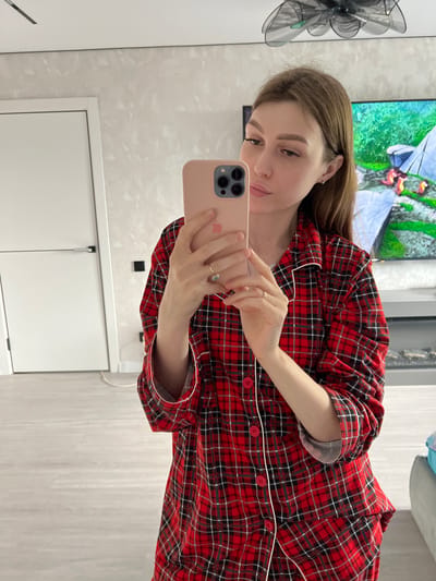A young woman is captured in a mirror selfie, standing indoors, likely in a home setting. She is seen from the chest up, with her head tilted slightly as she looks towards her reflection in the mirror, holding a light pink smartphone with multiple camera lenses. Her expression is calm and relaxed, with a subtle hint of a smile.

She has long, light brown hair and is wearing a long-sleeved pajama top or shirt featuring a red, black, and white plaid pattern, accented with white piping along the seams and red buttons down the front. Rings are visible on the fingers of both hands, which are holding the phone.

In the background, a large flat-screen television is mounted on the wall, displaying what appears to be an outdoor scene with green foliage and possibly some tents or structures. To the left of the woman, a decorative, dark-colored fan-like object with intricate patterns is mounted on the wall. The walls have a light, textured finish, and the flooring is also a light color, possibly wood or laminate. Below the TV, a white cabinet or storage unit is visible. No discernible text is present in the image. The overall setting suggests a comfortable, contemporary indoor environment.