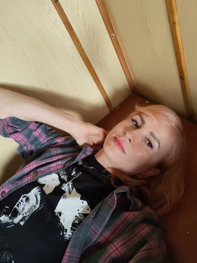A young woman with light brown hair, wearing a black t-shirt with a graphic print and an open plaid shirt, lies on her side. Her left arm is bent and rests behind her head, while her right arm is extended across her chest. She has full lips, dark eyebrows, and light-colored eyes. The background consists of light-colored wooden panels with wooden trim. The image does not provide any information about the location in the city of Kirillov, Russia.