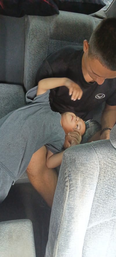 In a vehicle interior, two individuals are seated. The person in the foreground, a child, is lying on their back with their head resting on the lap of the second individual, an adult. The child is wearing a grey t-shirt and appears to be asleep or resting. The adult, wearing a black t-shirt, is leaning over the child, their hand gently resting on the child's arm or chest. The seats of the vehicle are upholstered in a grey fabric. The lighting suggests an indoor or shaded environment, typical of being inside a car. The image's content does not provide any information about the city of Kargat, Russia.