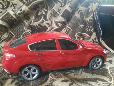 A red toy BMW X6 sits on a patterned fabric. The toy car is facing right, with its rear end visible. The fabric has a floral and geometric design in shades of brown, beige, and black. The car has black tinted windows, silver rims, and a BMW logo on the wheels. The car also has a fuel door on the rear passenger side. The background fabric appears to be a blanket or upholstery. There is no indication of the location in Zosna, Latvia.