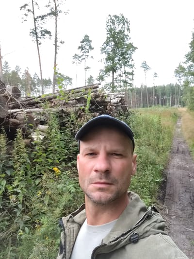 In the city of Tsaryuchski rural council, Belarus, a selfie captures a man wearing a blue baseball cap and a green jacket. He is standing in a forest with a pile of cut logs to his left and a dirt path to his right. The background is filled with trees and dense foliage. The lighting suggests an overcast day.