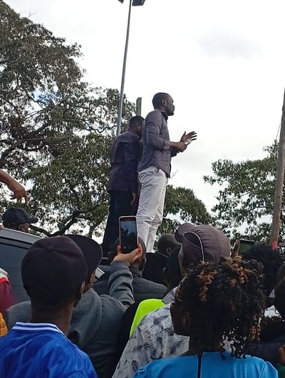 The image depicts an outdoor public gathering in Mukuyuni, Kenya, under an overcast sky. In the upper-central portion, a man wearing a dark patterned shirt and white trousers stands on the roof of a dark-colored vehicle, addressing an unseen audience. He gestures with his right hand open and holds a small, light-colored object in his left hand. A second man in a dark shirt stands partially visible behind the speaker. The foreground is occupied by the backs of multiple individuals in the crowd, looking towards the speakers. Several individuals are actively recording the event with smartphones; one phone, held by a hand with red nail polish, is prominently displayed in a vertical orientation, capturing the speaker on its screen. The environment includes mature green trees in the background and a tall metal pole with a light fixture extending upwards on the right side, indicative of a public space.