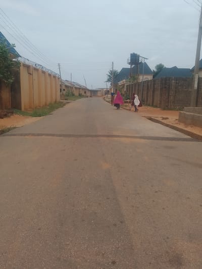 The image depicts a paved road in Gindi Nassarawa Kyarmi Galbi, Nigeria, under an overcast sky. The road is bordered by walls and buildings on both sides. On the left, a long, light-brown wall stretches into the distance, topped with barbed wire. Beyond this wall, glimpses of buildings with blue roofs are visible, along with some foliage. On the right side of the road, a rough, dark-grey stone wall lines the street. Further down the road, on the right, a black water tank stands on a metal structure atop a building with a dark, pitched roof. Three individuals are walking away from the camera on the right side of the road. Two are wearing flowing garments; one in pink and the other in white. The third individual, further ahead, is dressed in brown. The road itself is asphalt, showing some wear and tear with lighter-colored patches. The overall scene suggests a residential or semi-commercial area in a Nigerian town.