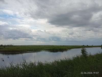 This is a wide-angle, full shot of a tranquil body of water, likely a river or a canal, bordered by lush green reeds and tall grass. The water reflects the overcast, cloudy sky, which dominates the upper half of the image. In the background, a flat, grassy landscape stretches to the horizon, punctuated by a few scattered buildings and utility poles. The scene is devoid of human presence or any discernible activity, conveying a sense of natural quietude. The image includes a timestamp in the bottom right corner, reading "22.06.2026 19:07", suggesting it was captured on June 22, 2026, at 7:07 PM. The location is identified as Sarytogay, Kazakhstan.