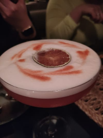 A shallow, wide-rimmed cocktail glass occupies the central foreground, containing a reddish-pink liquid beneath a thick, opaque white foam layer. A thin, circular slice of reddish-pulp citrus, likely a blood orange or grapefruit, rests centered on the foam. Irregular, reddish-orange streaks are present on the white foam, radiating outwards from the citrus slice. The cocktail is positioned on a dark, reflective surface, presumably a table. To the right, a blurred, light brown, woven texture is visible. The background is out of focus and dimly lit, revealing parts of two individuals. In the upper left, a blurred human hand with dark nail polish and a metallic accent near the cuticle is partially visible. In the upper right, a person wearing a blurred light green garment is discernible. The scene depicts an indoor social setting, likely a bar or restaurant, but the specific location within Bayreuth, Germany, is not identifiable from the visual evidence.