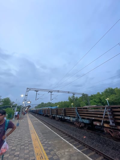 The image depicts a railway station platform in Sircaim, India, under a cloudy, twilight sky. Several people are visible on the platform, some appearing to be waiting or walking. A train, comprised of multiple flatbed carriages, is parked on the tracks alongside the platform. The platform itself is paved with interlocking bricks and marked with a yellow safety line. Overhead, complex electrical catenary systems for the train are prominent against the sky, with supporting structures and wires stretching across the frame. Trees and lush green foliage line the area beyond the train tracks, suggesting a rural or semi-urban setting. A digital display board with the time "01:20" is visible on a gantry. The overall mood is subdued, likely due to the overcast weather and the time of day.