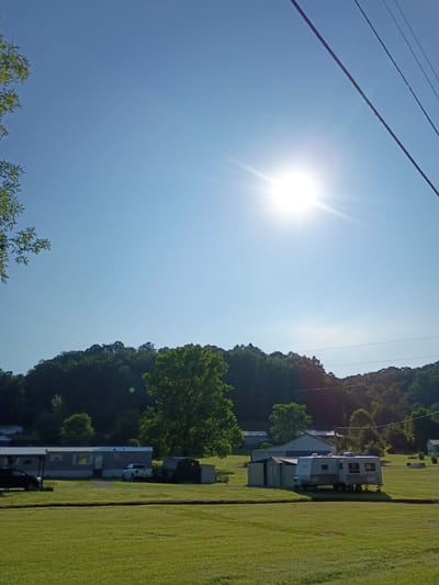 A bright sun shines in a clear blue sky over a rural landscape. In the foreground, a well-maintained lawn stretches out, bordered by a slight depression. In the middle ground, a long, light-colored trailer home with a dark roof is parked, with a dark-colored car to its left and a white pickup truck to its right. Adjacent to the trailer home is a smaller shed-like structure. Further to the right, a larger recreational vehicle is parked on the grass. Behind the structures, a line of trees forms a dense treeline that extends across the horizon. In the distance, rolling hills covered in trees rise, with power lines crisscrossing the sky. The image appears to be taken in a location resembling Berea, United States, given the rural setting.