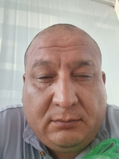 The image is a close-up, low-angle shot of a man's face, with the camera positioned below his chin, looking upwards. The man has closed eyes and is wearing a grey collared shirt. A portion of a bright green plastic bag is visible in the lower left of the frame. The background consists of a white, slatted surface, possibly blinds or a textured wall, suggesting an indoor setting. The lighting appears to be artificial and casts a soft glow on the man's face. The man's expression is neutral or serene due to his closed eyes. There is no visible text in the image. The overall impression is an informal, candid portrait, perhaps taken in a casual moment. The provided location context of Gulistan, Uzbekistan, does not offer specific visual cues within the image itself but might indicate a general geographical setting for the photograph.