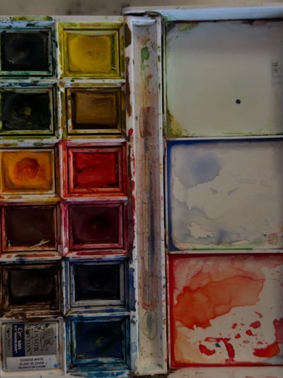 A well-used watercolor palette displays an array of colors in square compartments. The top row reveals yellow, brown, red, dark red, black, and blue. The bottom row shows black, green, yellow-orange, brown-red, and black. A Winsor & Newton "Cotman" watercolor tin of Chinese White sits at the end. The lid exhibits three large, stained mixing areas: one white with a small blue dot, a blue-tinged white, and a red-orange. Splatters and dried paint mark the tray, indicating frequent use. "mk11" is written on the inside of the lid.