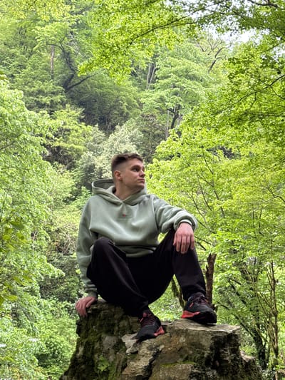 A young man is sitting on a moss-covered rock outcropping in Tsabal, Abkhazia, with a dense forest of green foliage as a backdrop. He is wearing a light green hoodie, dark pants, and dark sneakers with red accents. He is looking upwards, seemingly towards the sky or treetops.
