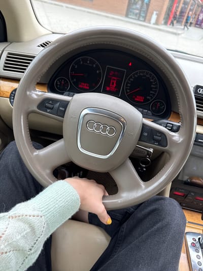 The image shows a close-up of the interior of an Audi A4 (B7) sedan, viewed from the driver's perspective. The steering wheel, dashboard, and part of the center console are visible. A person's hand, wearing a light blue knitted sweater, is resting on the steering wheel. The dashboard displays the time as 14:27 and the odometer reading as 15333. The car is likely stationary, as the gear selector is visible in the "P" (Park) position. The exterior environment, seen through the windshield, suggests an urban setting with a brick building and shops. The lighting indicates it is daytime. The specific location within Yartsevo, Russia, cannot be determined from the visual information provided.