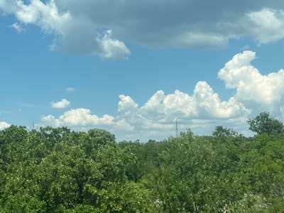 The image depicts a view of lush green trees under a partly cloudy blue sky. In the distance, a tall, slender communication tower is visible, rising above the dense foliage. The trees are full and leafy, suggesting a temperate climate and likely spring or summer. The clouds are varied, with some appearing fluffy and white, while others are darker and more diffused, hinting at potential weather changes. The scene appears to be an outdoor, natural environment, possibly on the outskirts of a town or city, given the presence of the tower. The exact location within the city of Vilusi, Montenegro cannot be determined from the image alone, as it lacks any specific landmarks or identifying features.
