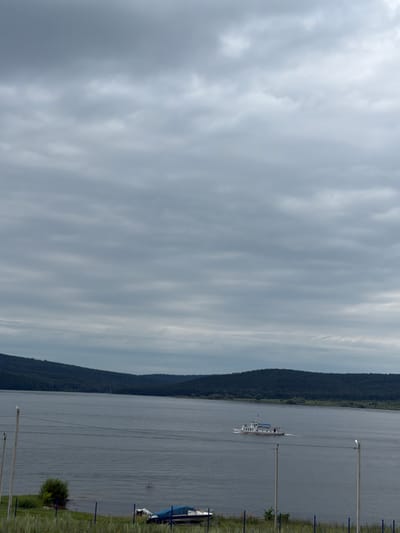 A wide, gray river dominates the lower half of the frame, reflecting an overcast sky filled with textured clouds. On the far bank of the river, a dark, densely forested hillside slopes upwards, its peak shrouded in the hazy atmosphere. In the middle distance, a white passenger boat with blue accents travels from left to right, leaving a wake on the water. The foreground features a grassy embankment with several utility poles and wires running horizontally across the scene. On the embankment, a blue tarp covers at least one boat. The location is the city of Sergeevka, Russia.