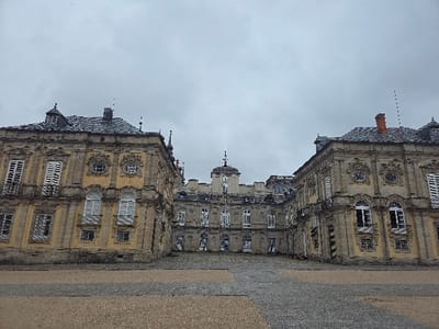 The image displays the Royal Palace of La Granja de San Ildefonso in the city of Real Sitio de San Ildefonso, Spain, viewed from its primary courtyard under an overcast sky. The scene is dominated by the palace's U-shaped architectural complex, comprising a central building flanked by symmetrical wings. Each wing presents a multi-story facade of light ochre plaster with prominent stone quoins and decorative surrounds for numerous windows. Windows are predominantly rectangular with white frames and closed white louvered shutters, while some circular or oval windows feature ornate stone detailing. The roofs are dark-tiled, exhibiting several chimneys and decorative spires. The central building, visible at the rear of the courtyard, features a lighter, uniform stone finish and is crowned by a central clock tower with a domed spire. The foreground comprises a large, open courtyard paved with a combination of lighter, possibly gravel, material in the immediate foreground, transitioning to darker, patterned stone or cobblestone closer to the palace entrance. No individuals or specific objects are discernible within the courtyard.