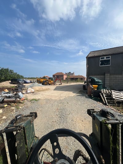 The image depicts an outdoor, unpaved yard or construction site located in Skipsea, United Kingdom, captured on a clear day with a blue sky and scattered white clouds. The perspective is from the operator's seat of a vehicle, showing a portion of a black steering wheel and dark green machinery components in the immediate foreground.
The midground features a large area covered in gravel and dirt. To the left, a substantial pile of assorted scrap materials, including metal and discarded objects, is present. Behind the scrap, a dark-colored minivan, a white hatchback, and a blue sedan are parked. Adjacent to these vehicles and the scrap, two distinct piles of aggregates—one of sand and one of grey gravel—are visible, along with a wooden pallet stacked with multiple white bags. Further into the midground, a yellow JCB-style backhoe loader is positioned, with a smaller red mini-excavator situated closer to the viewer.
To the right side of the scene, a two-story, dark grey building with a dark tiled roof features a single white-framed window on its upper level. Against the building's exterior, a large dark green storage tank with a yellow top, an orange gas cylinder, and a stack of wooden pallets are arranged.
In the background, a cluster of reddish-brown brick buildings with tiled roofs is visible. No individuals are present in the scene. All objects are static; no actions or interactions are depicted.