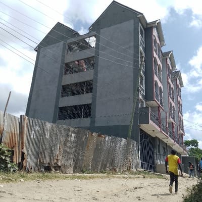 The scene depicts an urban environment in Ongata Rongai ward, Kenya. A multi-story residential building dominates the frame, viewed from a slightly low angle. The building is composed of at least two connected sections: the left section is five stories high, dark gray, with multiple rectangular windows fitted with dark security grilles, and visible laundry hanging on upper balconies. The right section, also five stories, is painted a lighter gray with pink/peach arched facades and black metal balcony railings. A dark metal gate is present at ground level on the right section.

In the foreground, an unpaved dirt road, strewn with small stones, slopes gently upwards to the right. A rusty, corrugated metal fence, appearing worn and partially collapsed, runs along the left side of the road, separating it from the building's property. Sparse green vegetation is visible on the far left behind the fence and along the roadside.

A male individual is walking away from the camera on the dirt road in the mid-ground, wearing a yellow short-sleeved top with red sleeves and dark trousers. Several other smaller figures are discernible further down the road to the right. A wooden utility pole supporting multiple overhead power or communication lines stands vertically in the mid-ground, slightly right of center. The sky is partly cloudy with white and gray clouds. The scene is illuminated by natural daylight.