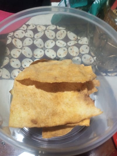 The image displays a clear plastic, round container holding a stack of approximately five to seven irregularly shaped, thin, light brown food items, consistent with fried or baked crackers. These items exhibit a slightly textured surface with some darker brown scorch marks or seasoning specks. The container sits on a dark brown textile surface patterned with repeating light beige, oval shapes, each containing a small dark brown cross or asterisk motif. The background beyond the container is out of focus, revealing indistinct dark and green elements on the left and right, suggesting an indoor setting. The specific location within the city of Buôn Ma Thuột, Vietnam, is not ascertainable from the visual information provided. No individuals are present in the image, and no specific actions or interactions are depicted.