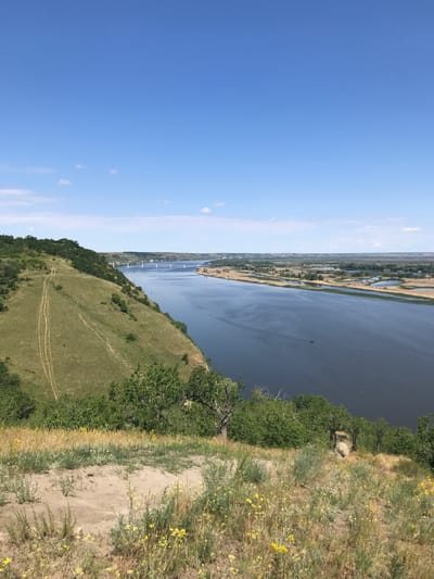 This is a high-angle, wide shot of a vast river bordered by a grassy, treed hill on the left and a flat landscape on the right. The sky is a clear, bright blue with a few scattered white clouds. The river dominates the center of the image, its dark water reflecting the sky. In the distance, a long bridge with white supports spans the river. The left side of the image features a steep, green hill with several dirt paths winding up its side. Lush green trees and vegetation cover the top of the hill and extend down its slopes. The foreground consists of dry, yellowed grass and scattered yellow wildflowers, with a patch of sandy earth. The right side of the image shows a flat, distant landscape with more vegetation and bodies of water, likely wetlands or oxbow lakes, along the riverbank. The scene is serene and natural, suggesting a rural or undeveloped area. This view is likely from the Mamaev Kurgan height in Kalach-na-Donu, Russia, overlooking the confluence of the Don and Volga rivers, possibly near the dam of the Volga-Don Canal.