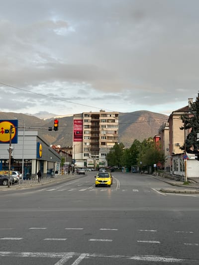 This image captures a daytime urban street scene in Usoika, Bulgaria, under an overcast sky. The photograph is taken from a slightly elevated perspective, looking down a street that stretches from left to right, with mountains forming a dramatic backdrop.

In the foreground, a multi-lane asphalt road dominates, marked with white dashed lines indicating traffic lanes and solid white lines along the shoulder. A yellow taxi cab, possibly a Hyundai i20, is visible driving on the right side of the road, heading towards the viewer. Pedestrians can be seen on the sidewalks, particularly near the modern, gray-fronted Lidl supermarket building on the left side of the street.

Along the street, various buildings line both sides. On the left, beyond the Lidl, older residential or commercial buildings with multiple windows are visible. A prominent red billboard on one of these buildings displays Cyrillic text: "ДОВЕРЕТЕ СЕ НА МНЕНИЕТО НА НАШИТЕ КЛИЕНТИ И ИЗБЕРЕТЕ КАЧЕСТВОТО НА JYSK" (Trust the opinion of our clients and choose the quality of JYSK). Traffic lights on the left side of the road show both red and yellow illuminated, suggesting an impending light change or a specific signal. Other street furniture includes lamp posts and various signs, including a red circular "No Entry" sign and a yellow signpost displaying the Lidl logo.

The background is dominated by large, forested mountains that rise behind the urban landscape. The sky is largely covered by uniform gray clouds, indicating an overcast day with no direct sunlight. The overall atmosphere is that of a typical, active town or city street.