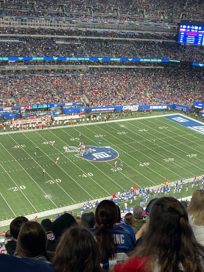 The image displays an American football game in progress at MetLife Stadium in East Rutherford, New Jersey, United States. The scene is viewed from an elevated seating position within the stadium, looking down at the green artificial turf field and across multiple tiers of spectator seating.

The stadium is artificially illuminated, indicating a night or evening event. Thousands of spectators are present, filling the vast majority of the seats visible in the foreground and across the background tiers. Blue LED ribbon boards displaying advertisements ("CUTWATER REAL COCKTAILS. PERFECTLY MIXED.") and "METLIFE STADIUM" are visible along the upper rim of the lower bowl. A large digital scoreboard showing game information is present in the upper right portion of the image.

On the field, marked with white yard lines and numbers (e.g., -20, -30, -40, 50), two teams are engaged in an active play. The midfield prominently features the New York Giants' blue "NY" logo. One team is primarily dressed in blue and white uniforms, while the opposing team wears predominantly red and white uniforms. Players are clustered around the line of scrimmage, near the 40-yard line on the far side of the field. Additional uniformed players and staff occupy the sidelines. At least one game official in a black and white striped uniform is on the field.

In the immediate foreground, the backs of multiple individuals' heads are visible, partially obscuring the bottom edge of the playing field. One person in the foreground wears a blue jersey with the name "KING" visible on the back.