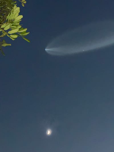 The image displays a twilight or nighttime sky above Manhattan Beach, United States. In the upper-center, a bright, pointed object emits a wide, arcing, luminous exhaust plume extending horizontally to the right across the sky. Below this phenomenon, a distinct crescent moon is visible in the lower-center of the frame. The upper-left corner features green foliage from a tree branch, partially entering the composition. No other subjects or actions are depicted.