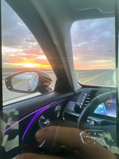 This is a first-person perspective shot from inside a car traveling on a highway at sunset. The exterior view shows a wide expanse of sky with colorful clouds, a vibrant orange sun low on the horizon, and a long, straight road stretching into the distance. The car's interior features a modern dashboard with a digital display showing a speed of 36 km/h and ambient purple lighting along the door panel. A hand is visible operating controls on the steering wheel. The side mirror reflects the road behind, showing the car's continued movement. The image is taken in an open, potentially rural or desert-like environment, with no identifiable city landmarks.
