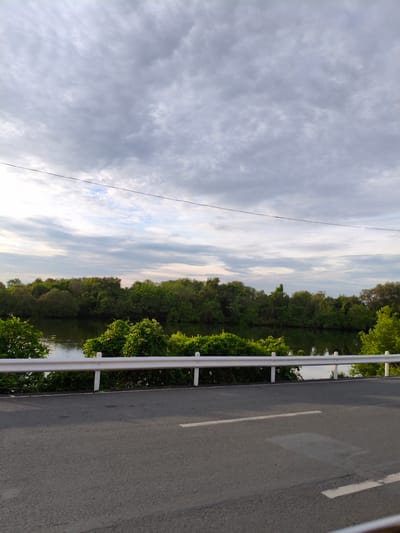 The image shows a view of a road adjacent to a body of water, likely a river or a cove, bordered by dense green foliage. The water reflects the overcast sky, which is filled with gray and white clouds. A white guardrail separates the road from the water and vegetation. The road itself is asphalt, with a white line marking its edge and another white line indicating a lane divider. The scene appears to be located in a natural or semi-natural area, possibly on the outskirts of a town or city. Given the request for the specific location, this scene is likely somewhere in Noveleta, Philippines, known for its coastal and riverine landscapes. No people are visible in the image, and there are no specific actions or interactions to describe. The overall impression is one of a quiet, scenic roadside environment.
