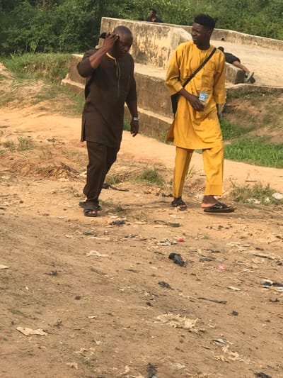 Two men stand outdoors on a dirt path. One man wears a yellow outfit and black shoulder bag, holding a small rectangular object. The second man wears a dark brown outfit and a watch, holding his hand to his ear. Both men wear sandals. The ground is littered with trash. Vegetation is visible in the background.