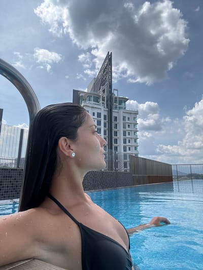 A young woman is shown relaxing in an outdoor swimming pool during the daytime, under a partially cloudy sky. She is depicted from the chest up, with her head tilted back and her gaze directed upwards. Her dark hair is wet, and water droplets glisten on her skin. She is wearing a black bikini top and a prominent stud earring in her right ear. Her right arm is partially submerged in the clear blue water, with her hand appearing to rest on the pool's edge. A chrome-plated handrail is visible in the immediate foreground.
In the background, tall, modern buildings rise against the sky. A distinctive white high-rise building with a complex structural element at its peak stands out, surrounded by other urban architecture. The setting suggests an elevated pool, possibly on a rooftop or a high floor, within an urban environment. The weather appears bright with significant cloud cover. The overall mood is one of relaxation and contemplation. The location context is Ampang Jaya Municipal Council, Malaysia. No visible text is present in the image.