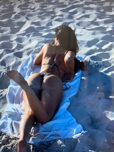 A person is lying on a white towel on a sandy beach. The person is wearing a camouflage bikini. The person's back is to the camera, and their legs are bent. The sand is rippled, indicating it is a windy day. The image does not provide enough information to determine the location in the city of Konstantinovka, Kyrgyzstan.