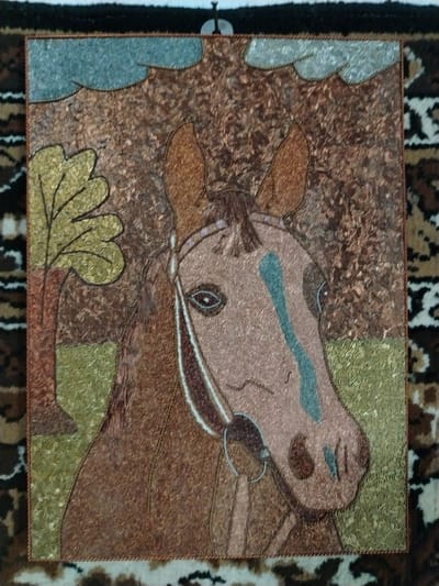 This is a square-shaped artwork of a horse's head and upper body, created with small, textured pieces of material, likely copper or metal, arranged to form a mosaic. The horse is depicted in profile, facing right. Its coat is primarily a reddish-brown hue, with a lighter, dusty rose color on its muzzle and a blue-green stripe down its forehead and nose. The horse wears a bridle with a metal bit. Its eyes are dark with a lighter pupil.
Behind the horse, the background suggests an outdoor scene. To the left, there is a stylized tree with a reddish-brown trunk and broad, yellowish-green leaves. The ground appears to be green and brown. Above the scene, there are stylized clouds in a light blue-gray color. The overall impression is of a pastoral or natural setting, possibly during the day.
The artwork is framed by a thin, twisted cord of the same material. It is displayed against a backdrop of a patterned rug with earthy tones and abstract designs, suggestive of a domestic setting or an exhibition space. There is no visible text in the image. The mosaic technique gives the artwork a rich, textured surface.
