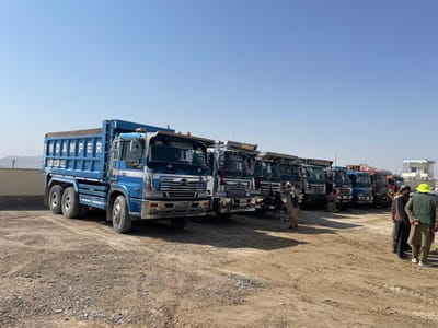 This image displays an outdoor truck lot in Pir Dust, Afghanistan, under a clear, pale blue sky. A linear arrangement of approximately 10-12 heavy-duty dump trucks is parked on an unpaved dirt and gravel surface. The prominent truck in the foreground is a blue and white tandem-axle dump truck, featuring "NDAMPER" in white capital letters on its blue dump bed and chromed front bumper and grille. Other dump trucks in the line vary in color, including darker blues and greens, with similar designs and visible modifications. On the far right, a group of 5-7 men, dressed in traditional attire, are standing and interacting; one man wears a bright yellow-green hat. Further left, a man stands near the front wheel of a dark truck, and another man stands further back, holding a pole-like object. In the background, distant mountains are visible to the left, and a small, light-colored building with a flat roof is present on the far right.