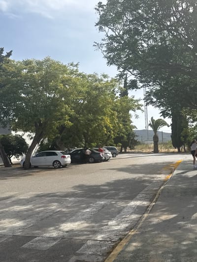 The image depicts a parking lot area in La Roda de Andalucía, Spain, with a street in the foreground marked with a crosswalk. Several cars are parked along the side of the road, including a white hatchback and a dark-colored sedan. Large trees with green foliage cast shadows across the pavement. In the background, a tall communication tower is visible against a hazy sky, with distant hills or mountains. A palm tree stands to the right of the tower. Two individuals are partially visible on the right side of the frame, one standing and another walking away. The scene appears to be a public outdoor space on a sunny day.