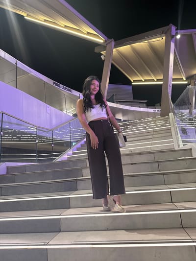 A young woman with long, dark, wavy hair is standing on a wide, outdoor staircase at night. She is wearing a white crop top with pearl embellishments and high-waisted, dark brown, wide-leg pants. Her shoes appear to be gold espadrille wedges. She is holding a light-colored purse with a chain strap. The staircase is made of concrete with integrated LED lights along the edges of each step, creating a luminous pathway. The background features a modern building with glass railings and a large, beige awning illuminated by warm yellow lights. A sign on the building reads "PURE CUISINE" and another sign says "100 MO." The environment is illuminated by artificial lighting, with streaks of purple light visible on the left side of the building. The overall scene suggests an outdoor dining or entertainment area in a city.